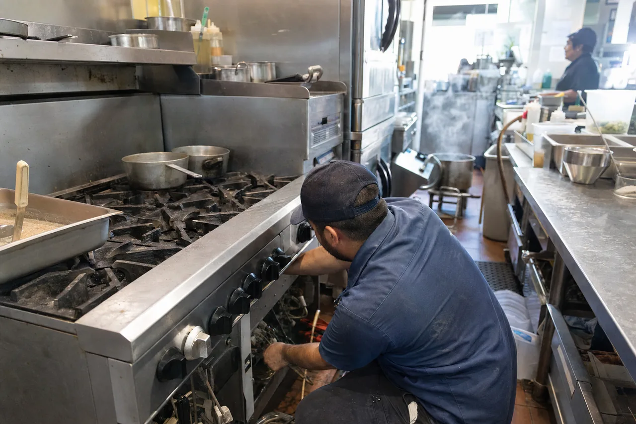A technician crouched at the control panel of a stainless gas range in a working kitchen, the knob row removed and pans still on the burners above